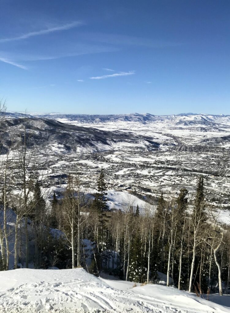 A view at the top of a ski mountain overlooking the town of Steamboat Springs Colorado. There are blue skies and there is snow covering the mountains and town.